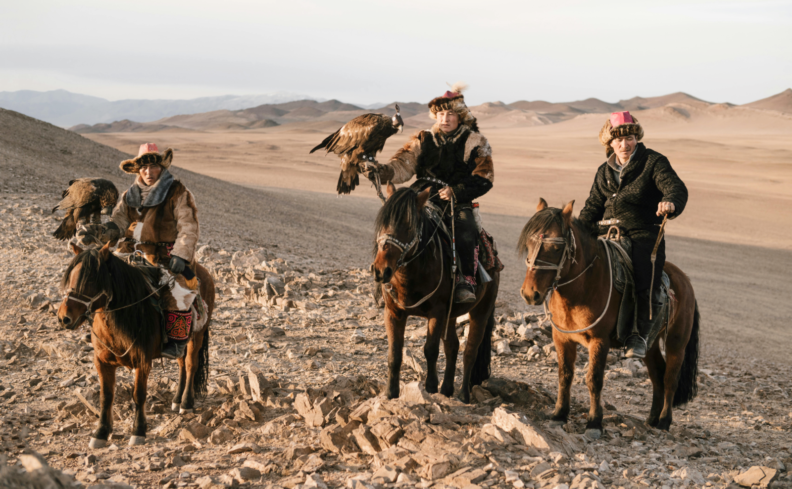 three mongolian falconers in traditional clothing sit on horseback while holding falcons on their wrists with the flat plains of the steppes behind them