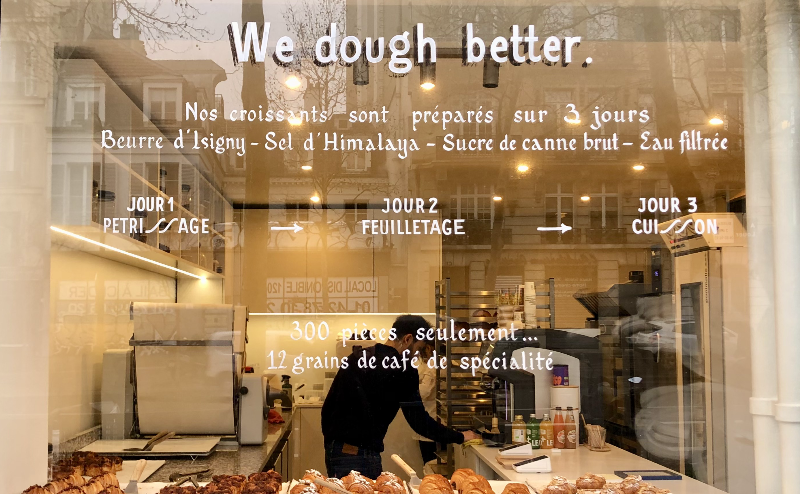 view through a large window of a man working in a bakery with a large tray of croissants in the foreground