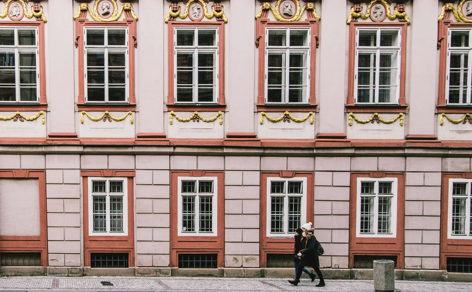 two women walking side by side in front of an ornate pink baroque building