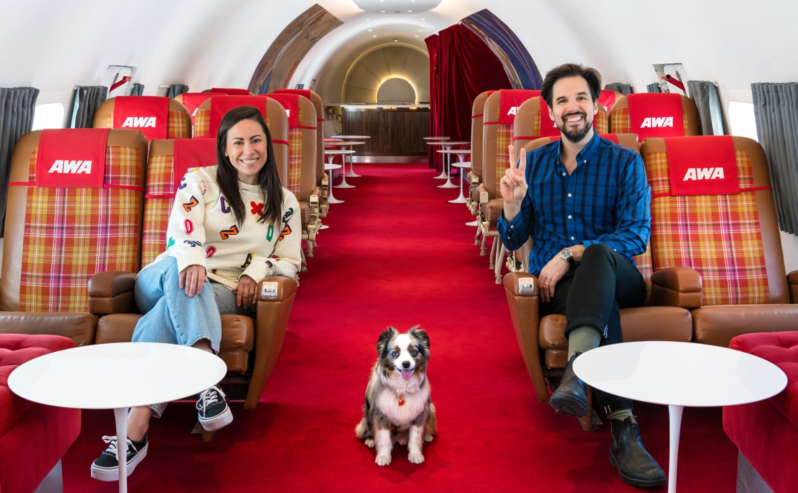 a woman, man, and small dog sitting in chairs in a vintage TWA airplane with red seats and carpet