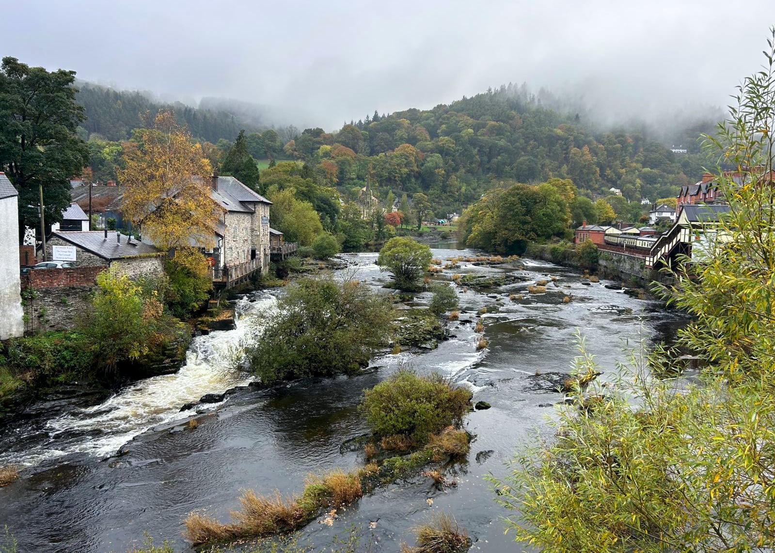 river running through a small town below a mountain covered in mist
