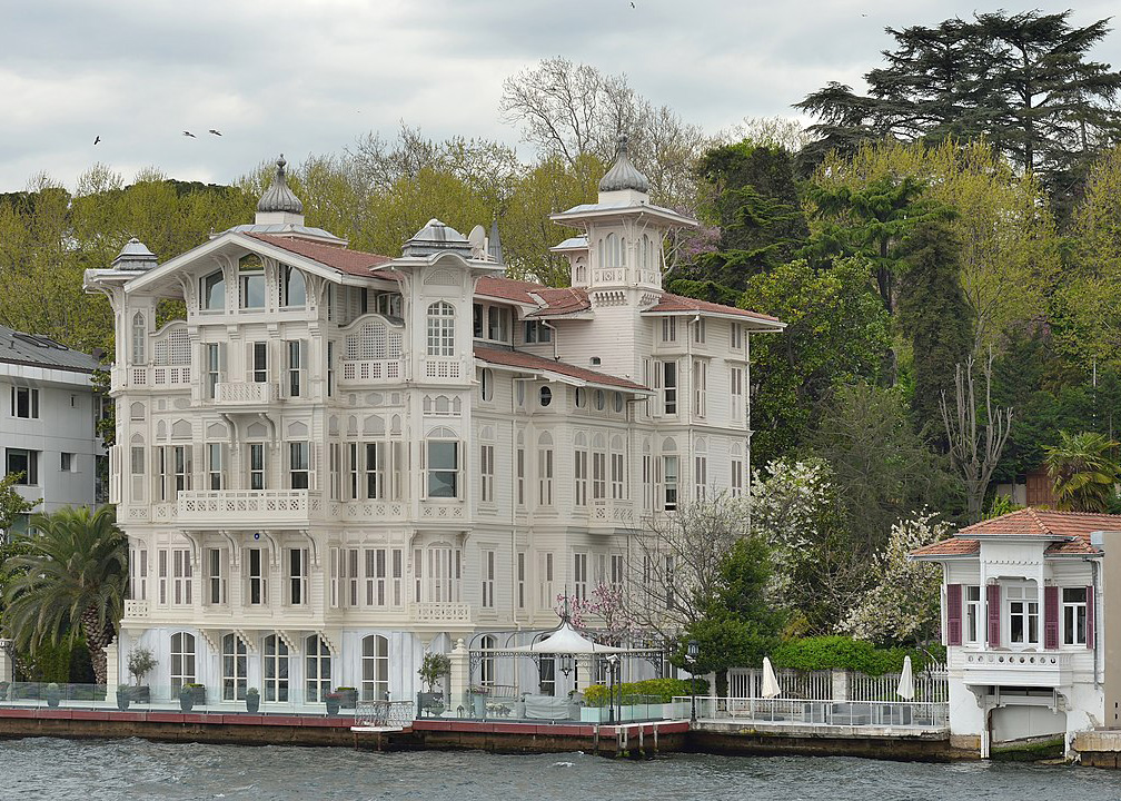 ornate white wooden house with four stories and small onion domes on the shore of the water