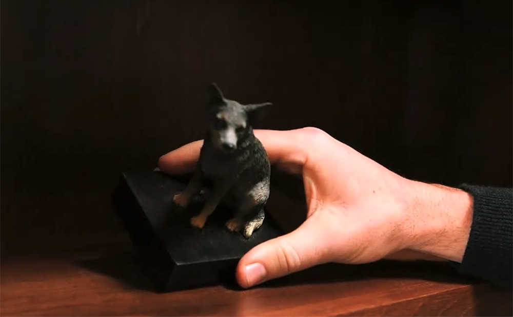 small black dog statue on a bookshelf being tipped by a human hand