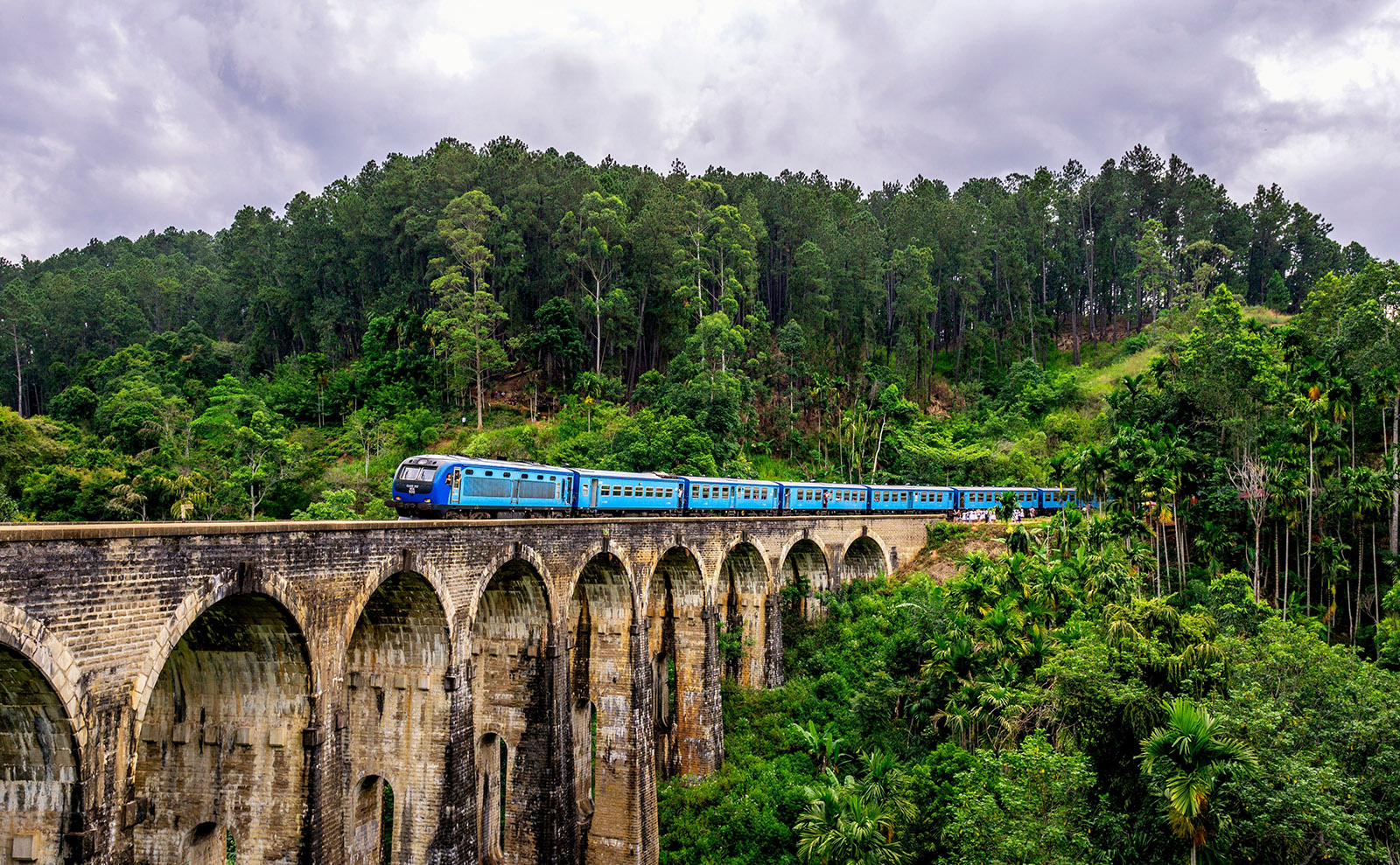 long blue train passing over a gray stone bridge in the jungle