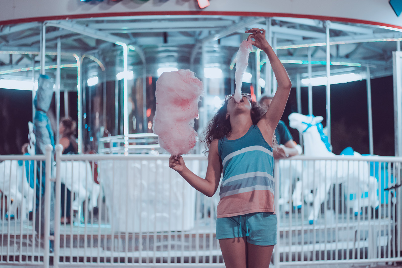 a girl in pink and blue shorts eating pink cotton candy in front of a blue merry-go-round