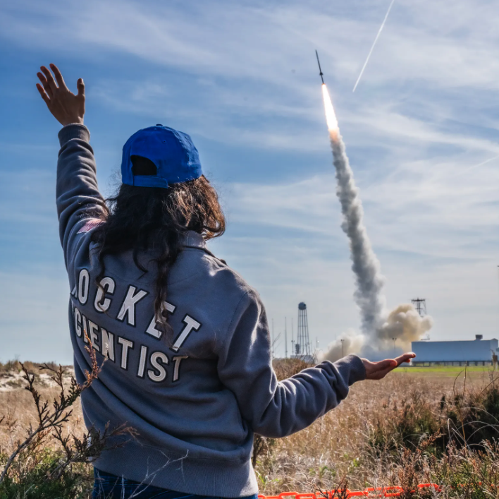 a woman wearing a jacket that says rocket scientist watches a rocket take off into the blue sky