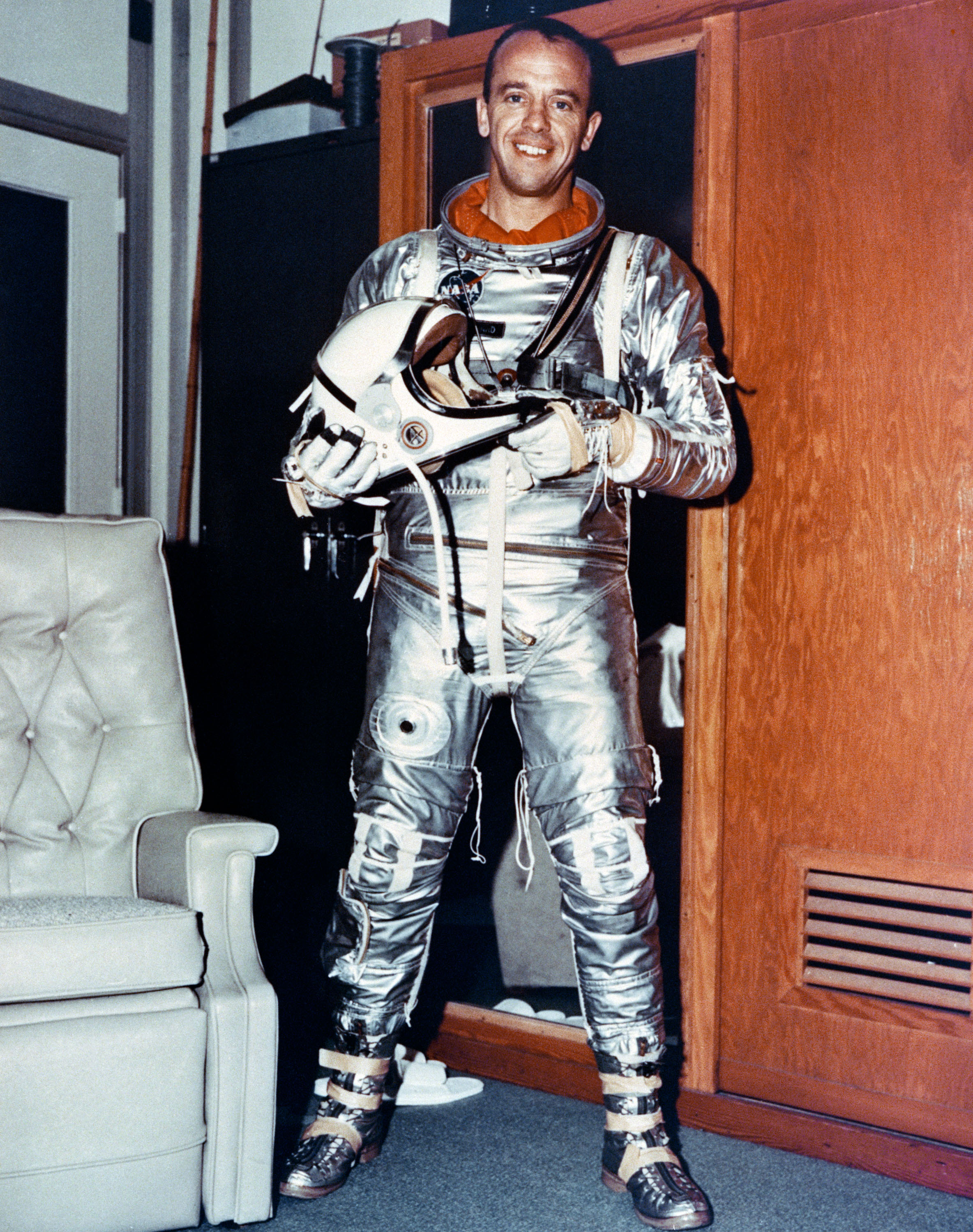 a man smiling wearing a metallic silver space suit in a room with 1970s wood paneling