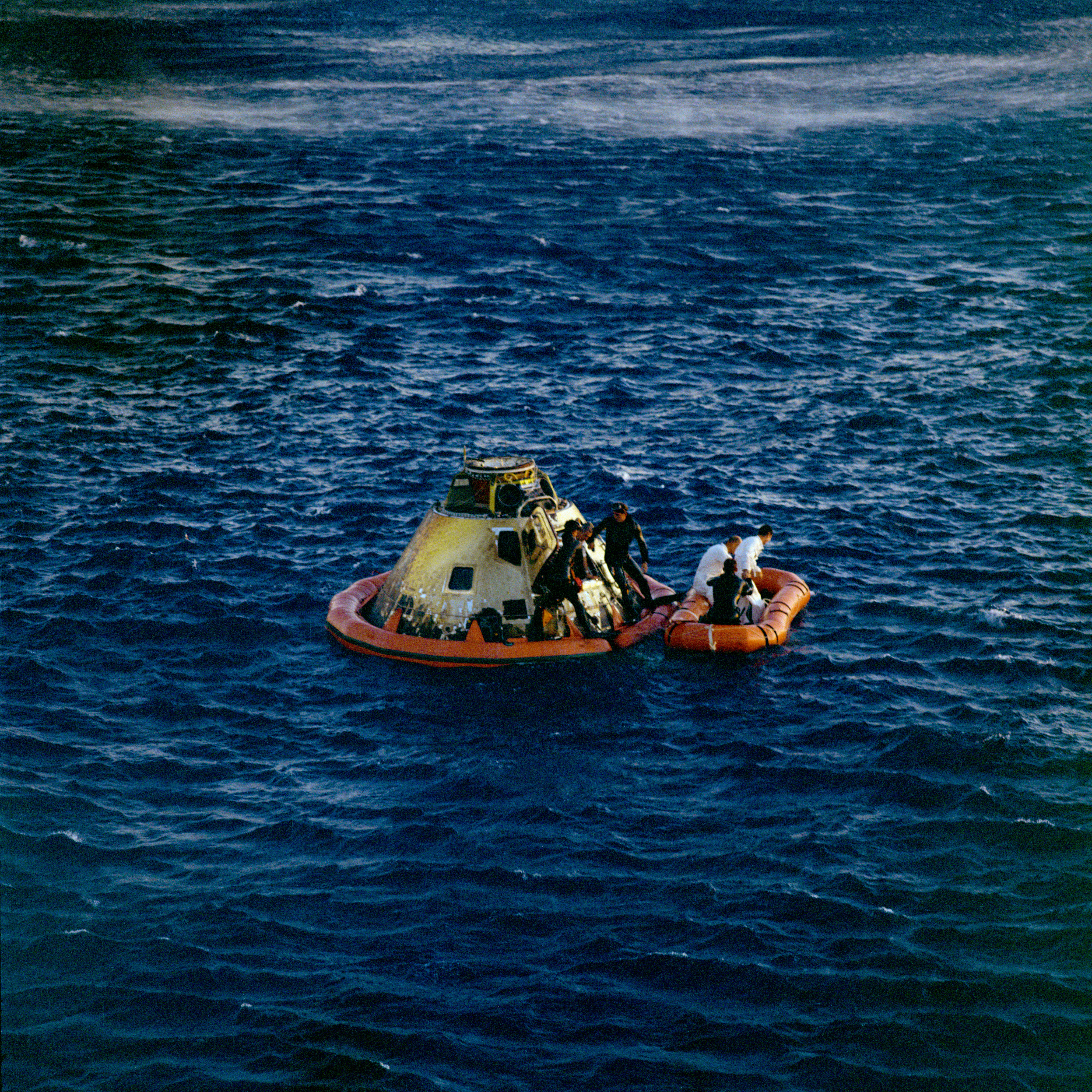 a space capsule floating in the ocean with men in orange rafts