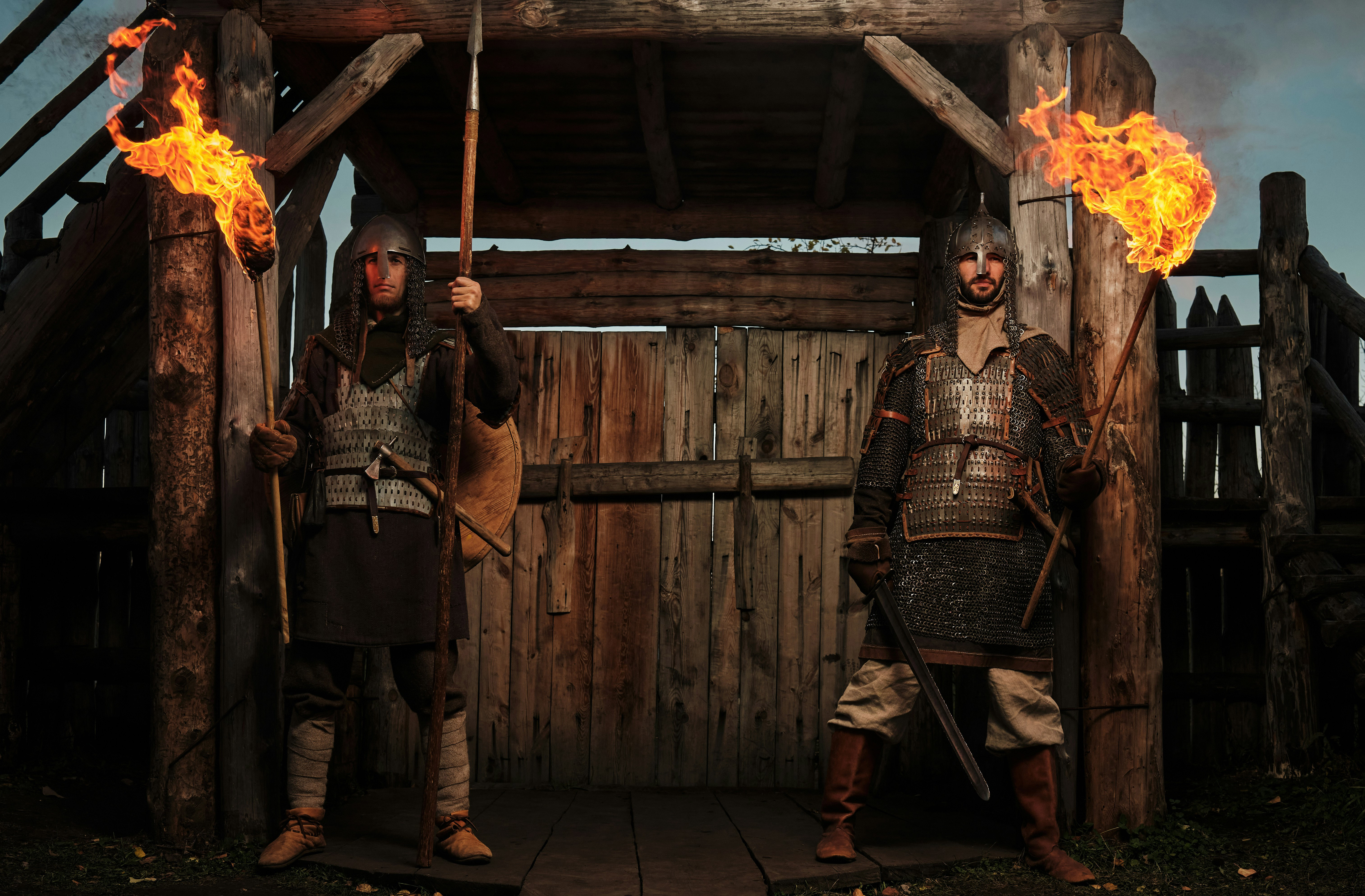 two men in viking helmets standing in front of a wooden house with flames behind them