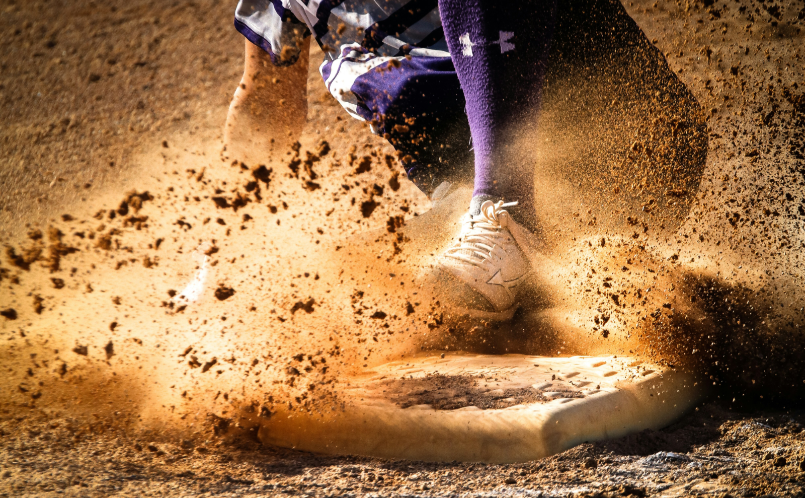 close-up of a person wearing purple pants sliding onto a base with sand-colored dirt flying in the air