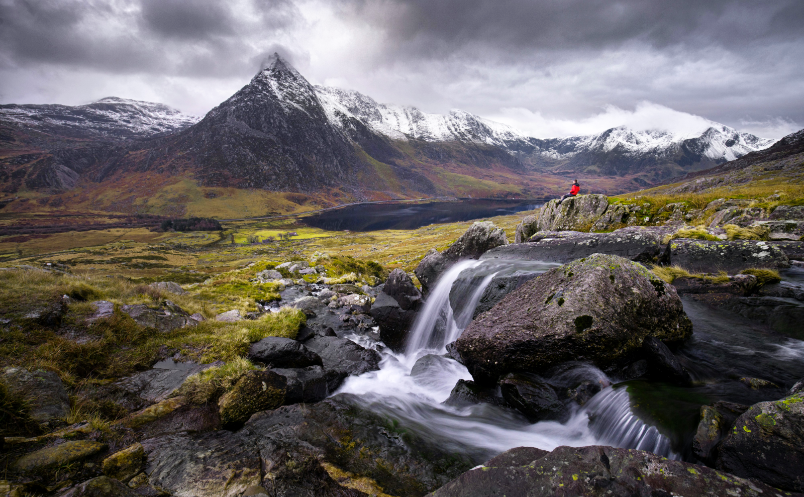 a gray foggy day with a waterfall in the foreground, a tall gray mountain topped with snow in the background, and a man sitting on a rock, wearing a red coat, in the middle