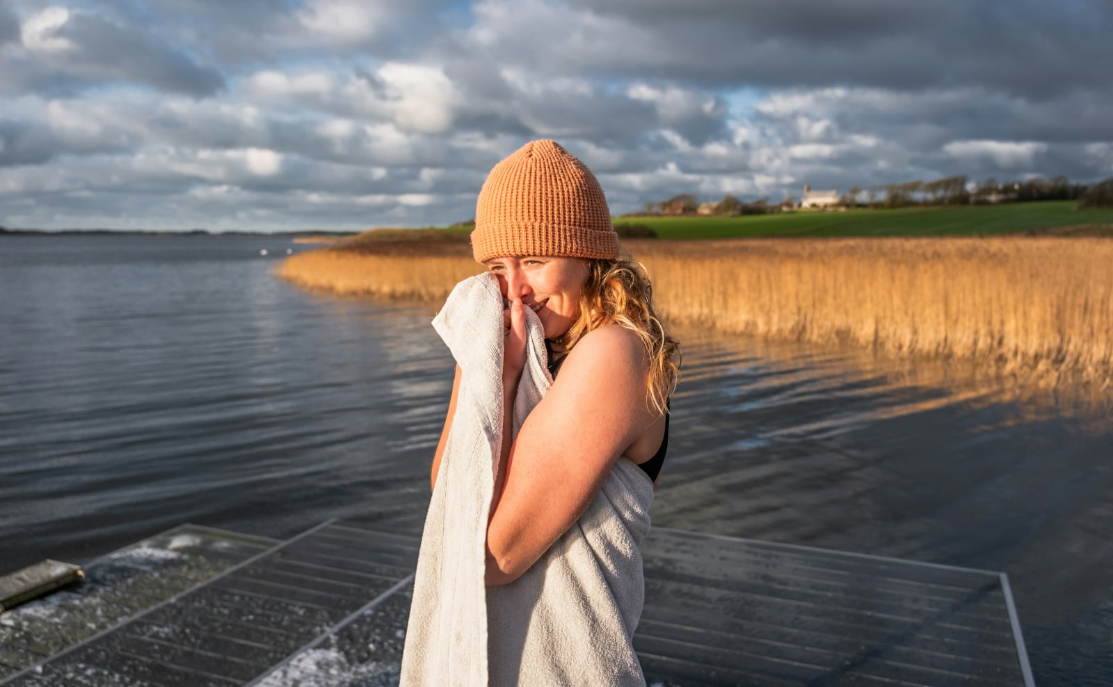 woman with blonde hair standing on a dock by the water drying her face with a towel