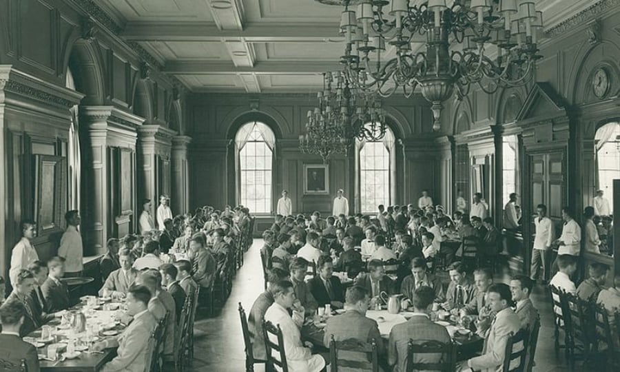 black and white photo of young men in a large dining room with crystal chandeliers and waiters