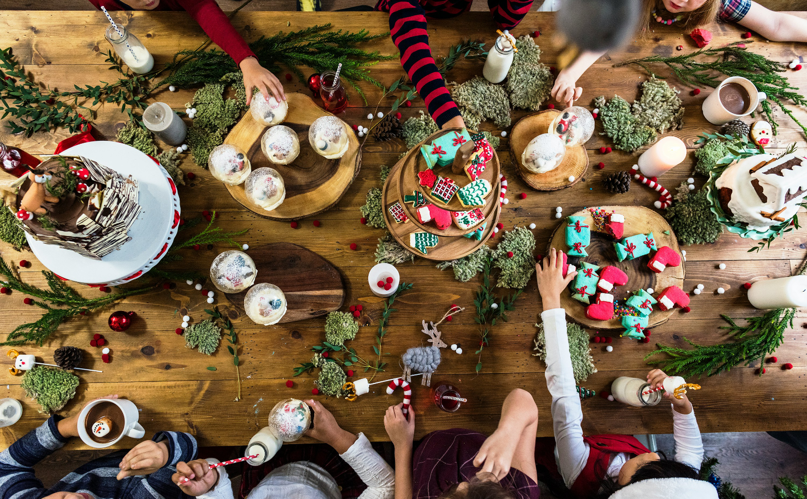 overhead view of people sitting around a wooden table decorated in red and green and topped with serving platters of food