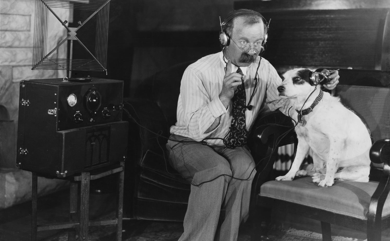 a retro photo of a man listening to the radio and holding earphones over his dog's ears