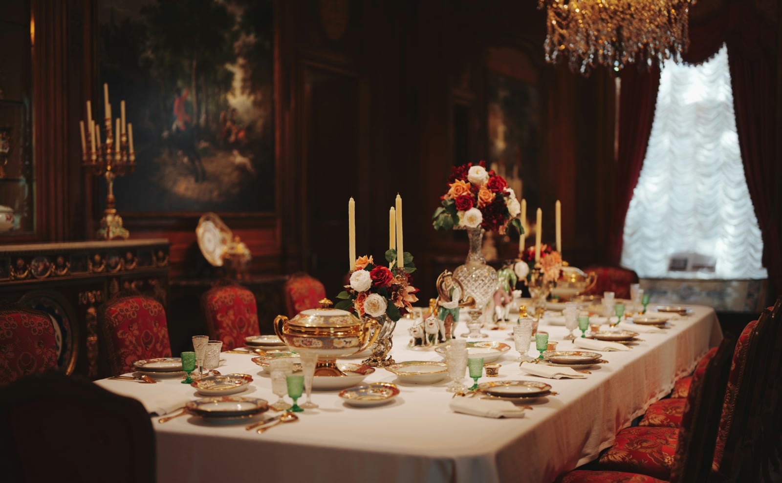 a long wooden dining table set with formal dinnerware and silver with a sparkling chandelier overhead