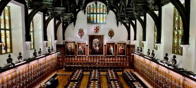 grand hall with tall ceilings and rows of long wooden tables with portraits on the walls