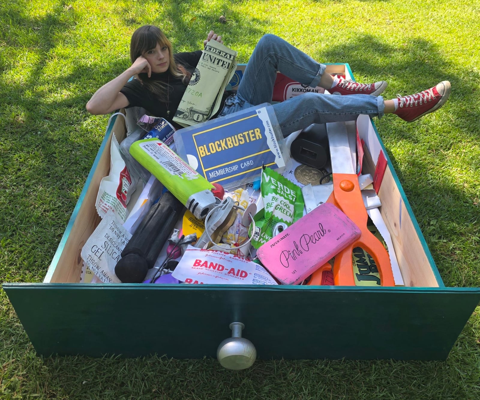 a giant junk drawer sculpture with scissors, keys, paper tickets and more with the artist sitting in the drawer