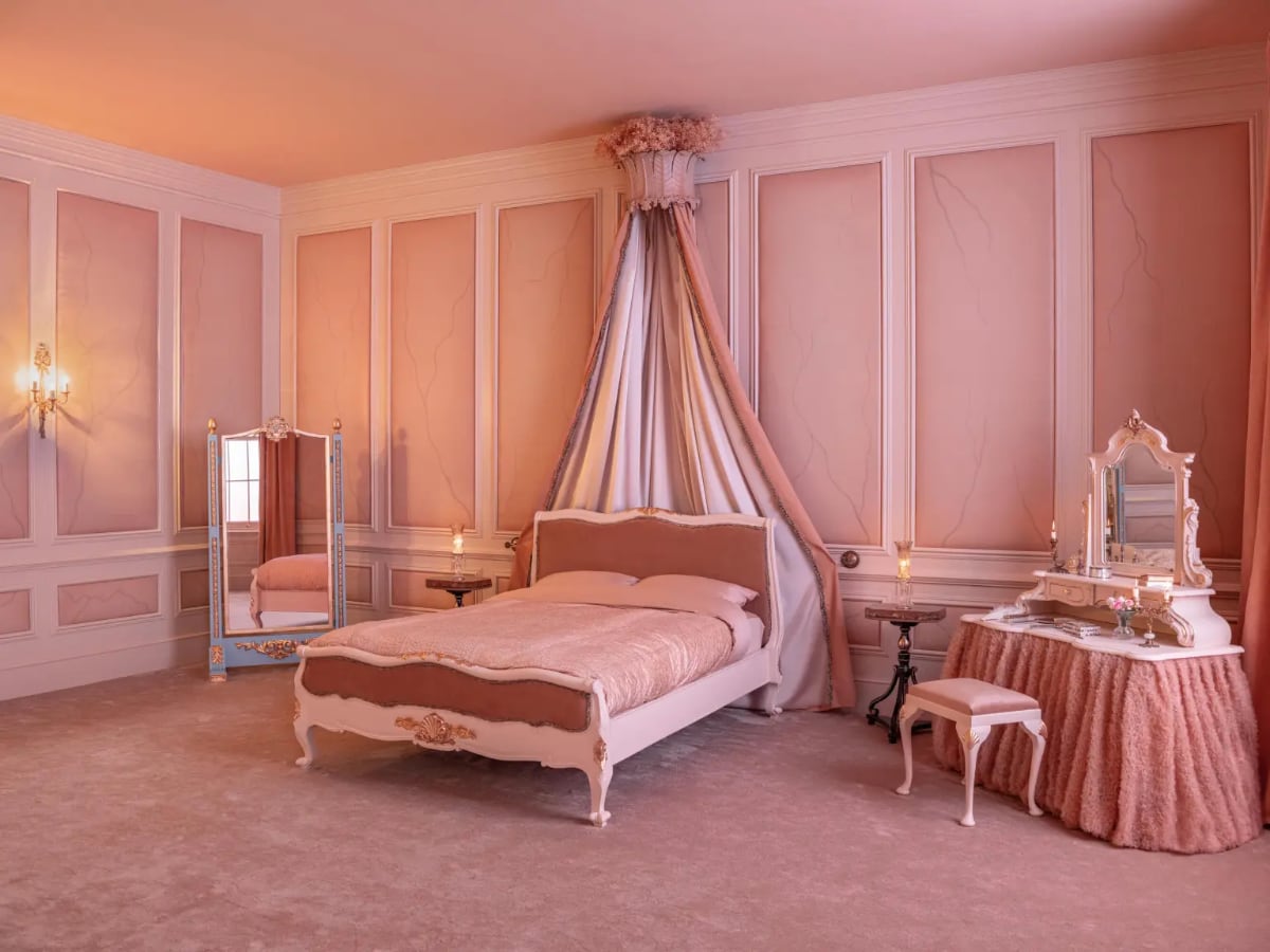 a bedroom with a big pink bed, pink walls, a canopy, and a dressing table with a mirror