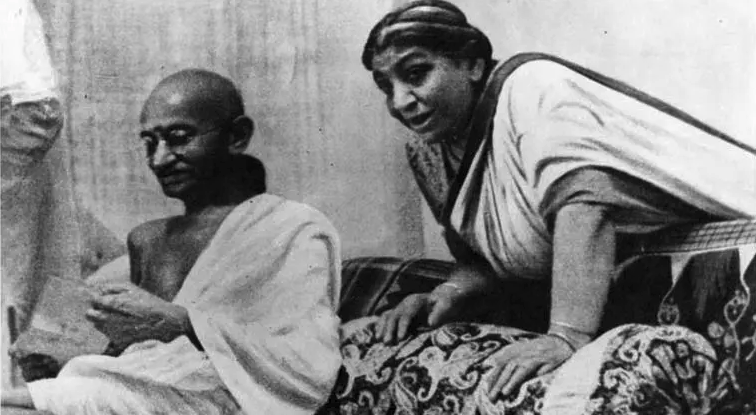 black and white photo of gandhi sitting on the left and sarojini naidu is leaning over to him from the right while wearing a sari