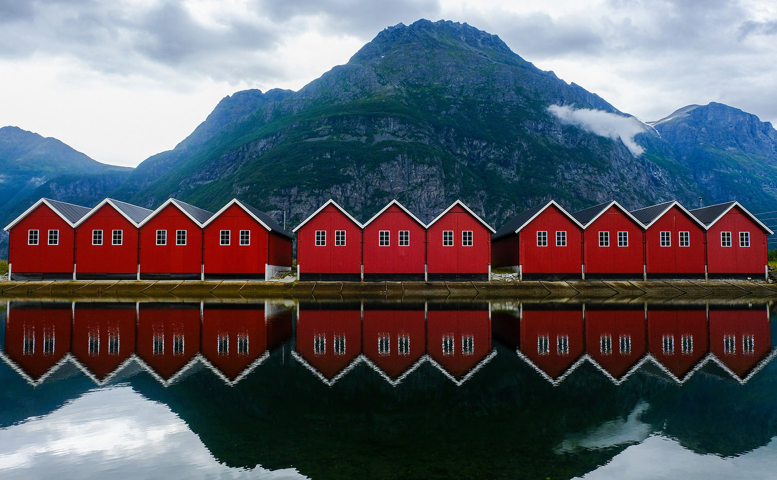  a row of red wooden houses with gray rocky mountains in the background and dark blue water in the foreground